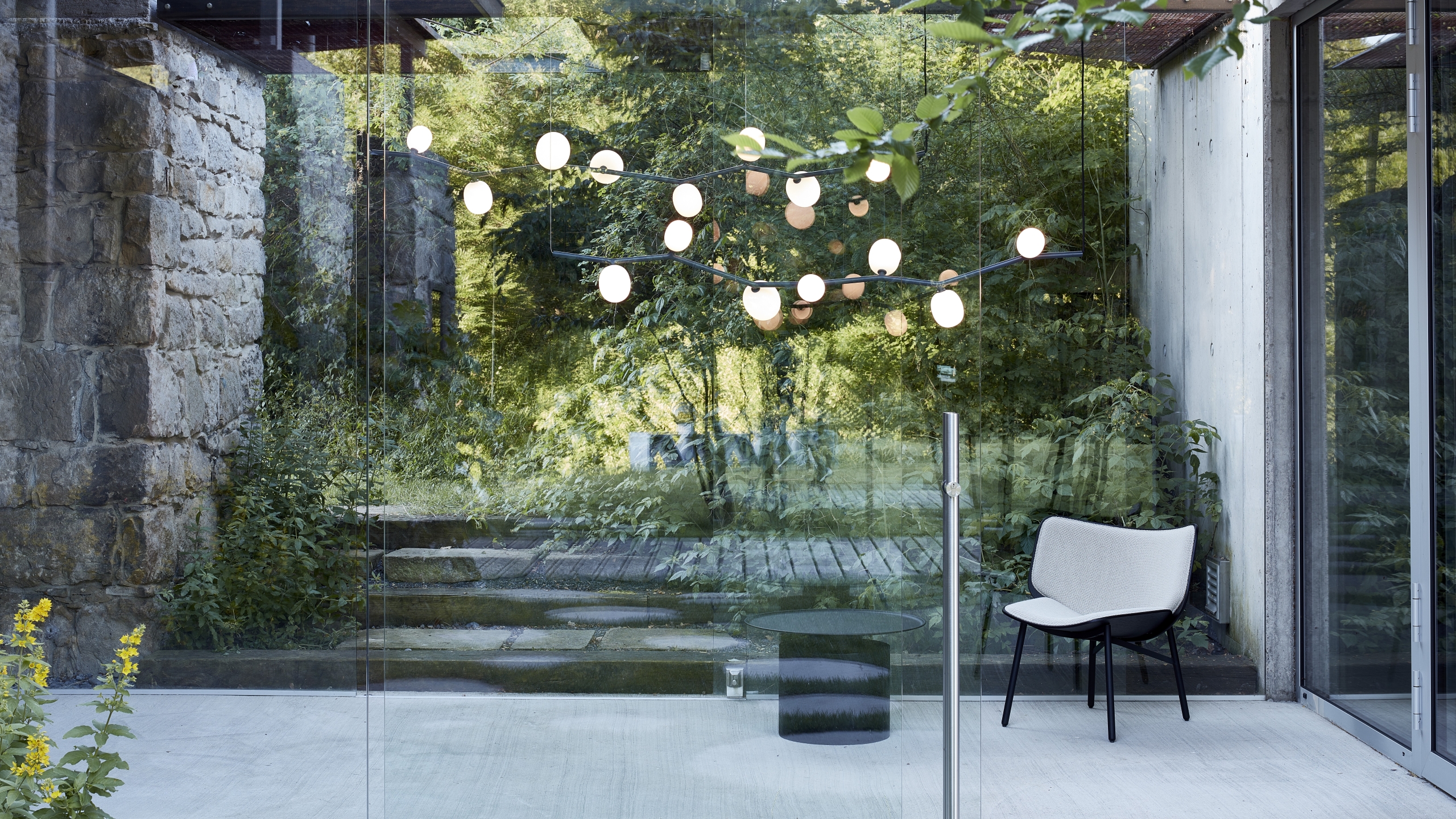 Brokis Ivy Horizontal chandeliers in a modern glass-walled room with a white chair, black table, and lush views.