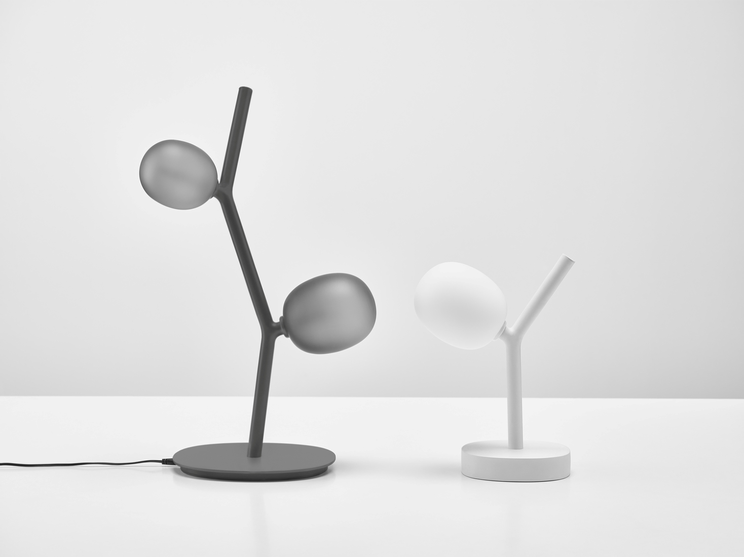 Brokis Ivy Table lamps - one dark grey with two glass ovals, one white with a single luminous oval - on white.