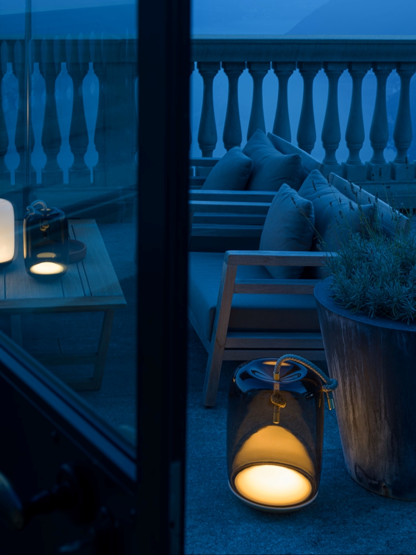 Elegant patio at dusk with a wooden sofa, grey cushios, potted plant, and Brokis Knot Battery lamps glowing warmly.