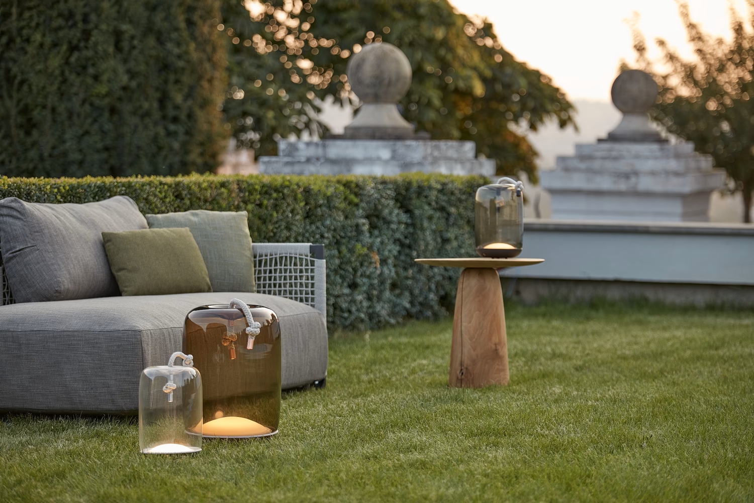 A luxe outdoor scene with a grey sofa, green cushions, and Brokis Knot Battery lanterns radiating a soft glow.