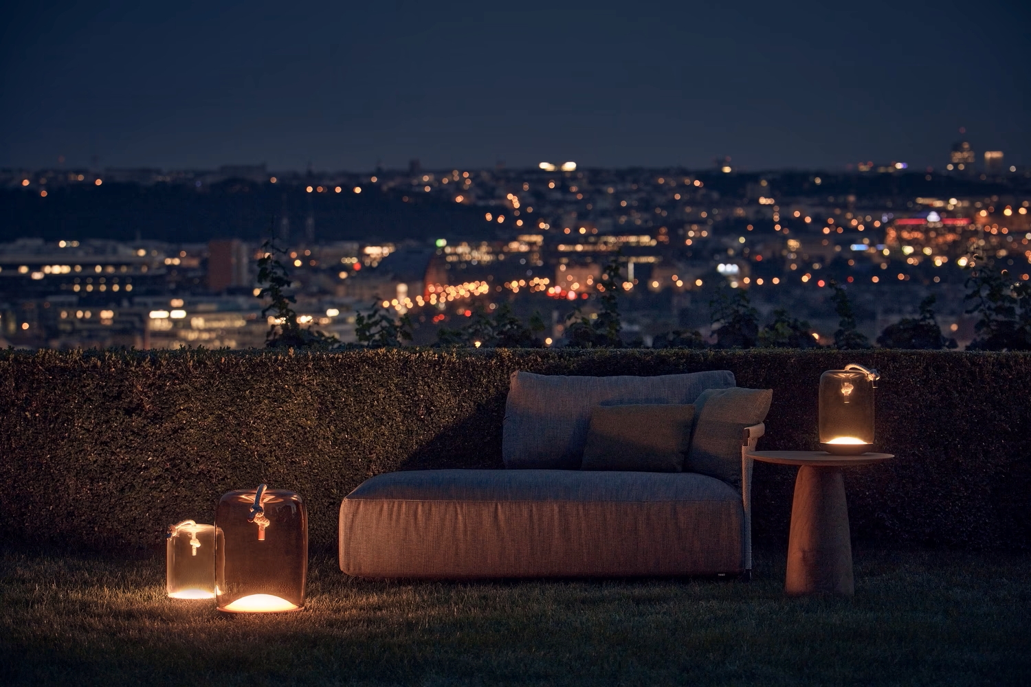 An outdoor lounge at night with plush seating, a wooden side table, and Brokis Knot Battery lanterns aglow.