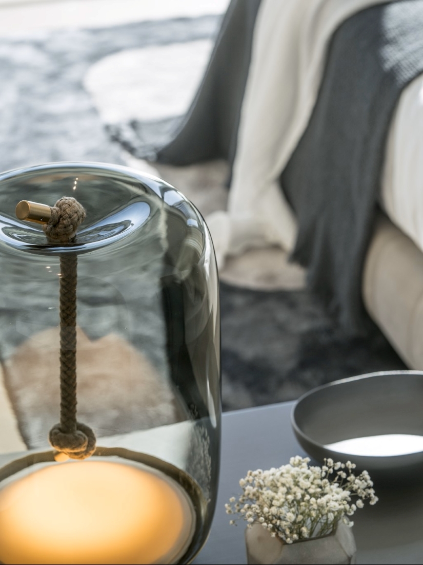 Brokis Knot Cilindro Table lamp with rope detail beside white blooms, framed by luxe bedding and a textured rug.