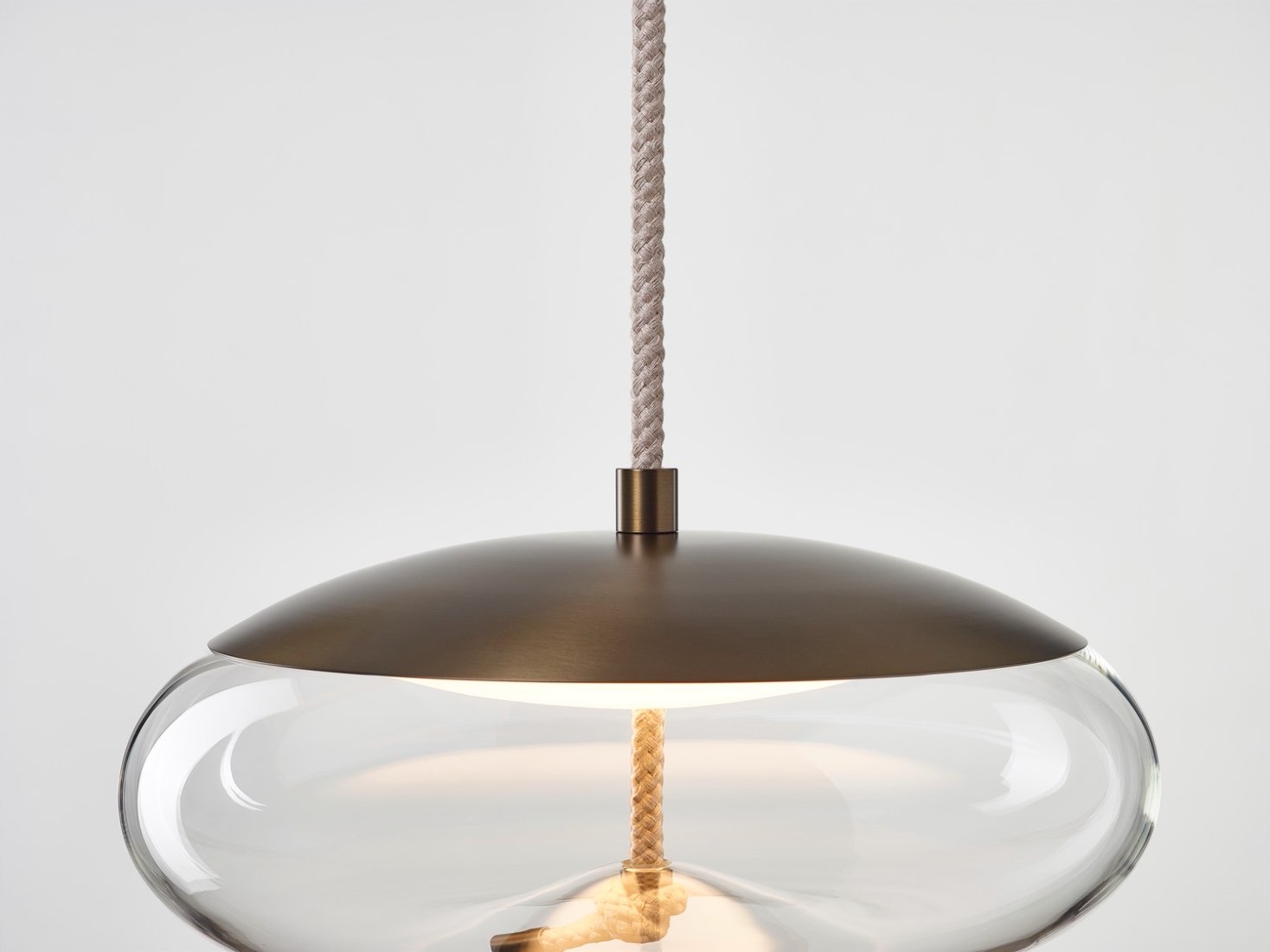 A Brokis Knot Disco Pendant with handblown clear glass and matt brass top hangs on textured rope, casting a golden glow.