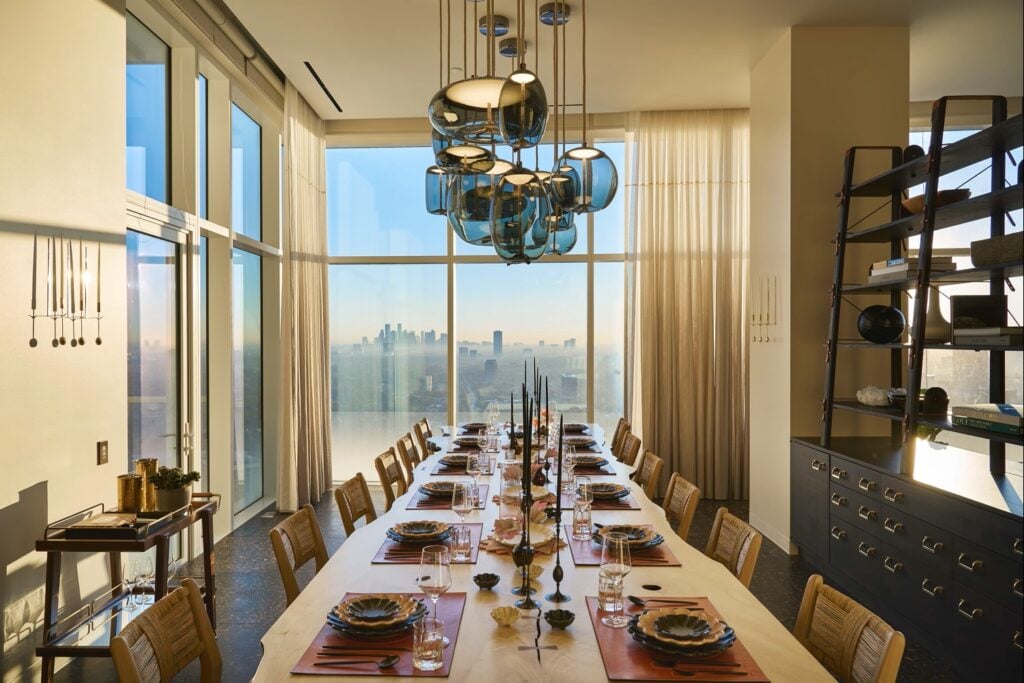 A dining room with a table for twelve, and Brokis Knot Pendants overhead; sunset skyline glows through large windows.