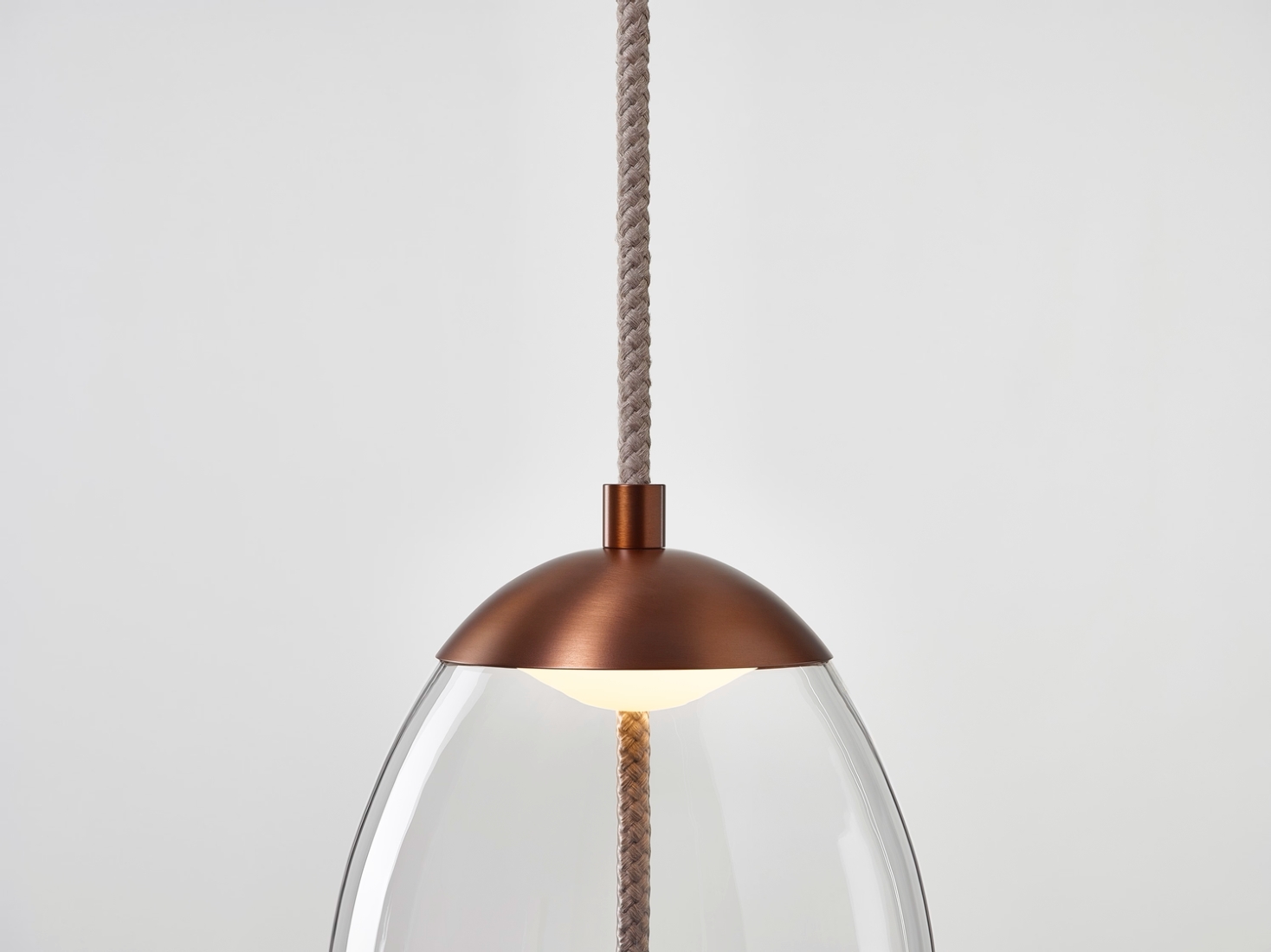 A Brokis Knot Uovo Pendant with a dome copper top, clear glass shade, and rope flex, casting a luxurious warm glow.