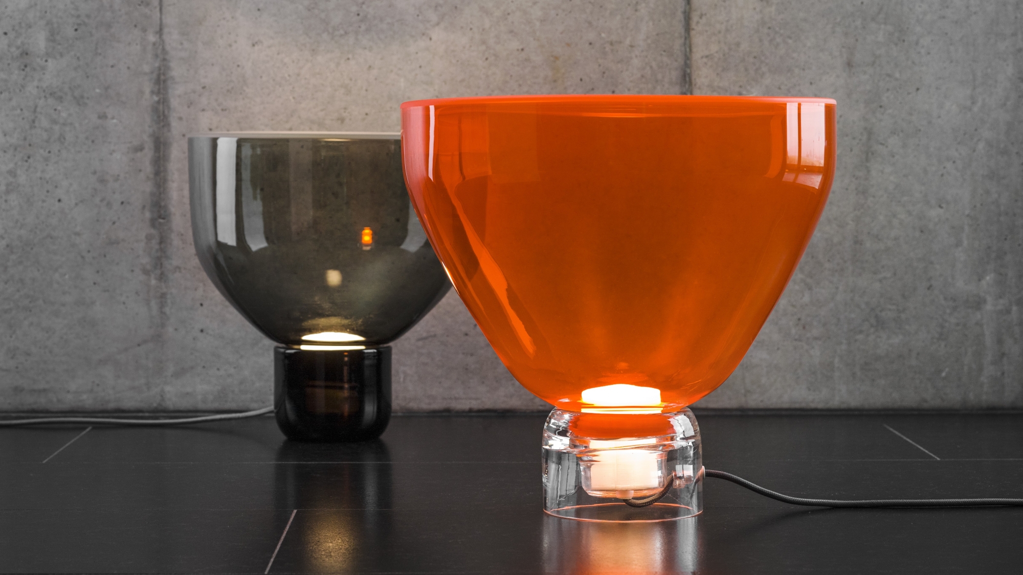 Brokis Lightline table lamps in orange glass and smoke grey cast a soft glow on a sleek black surface.