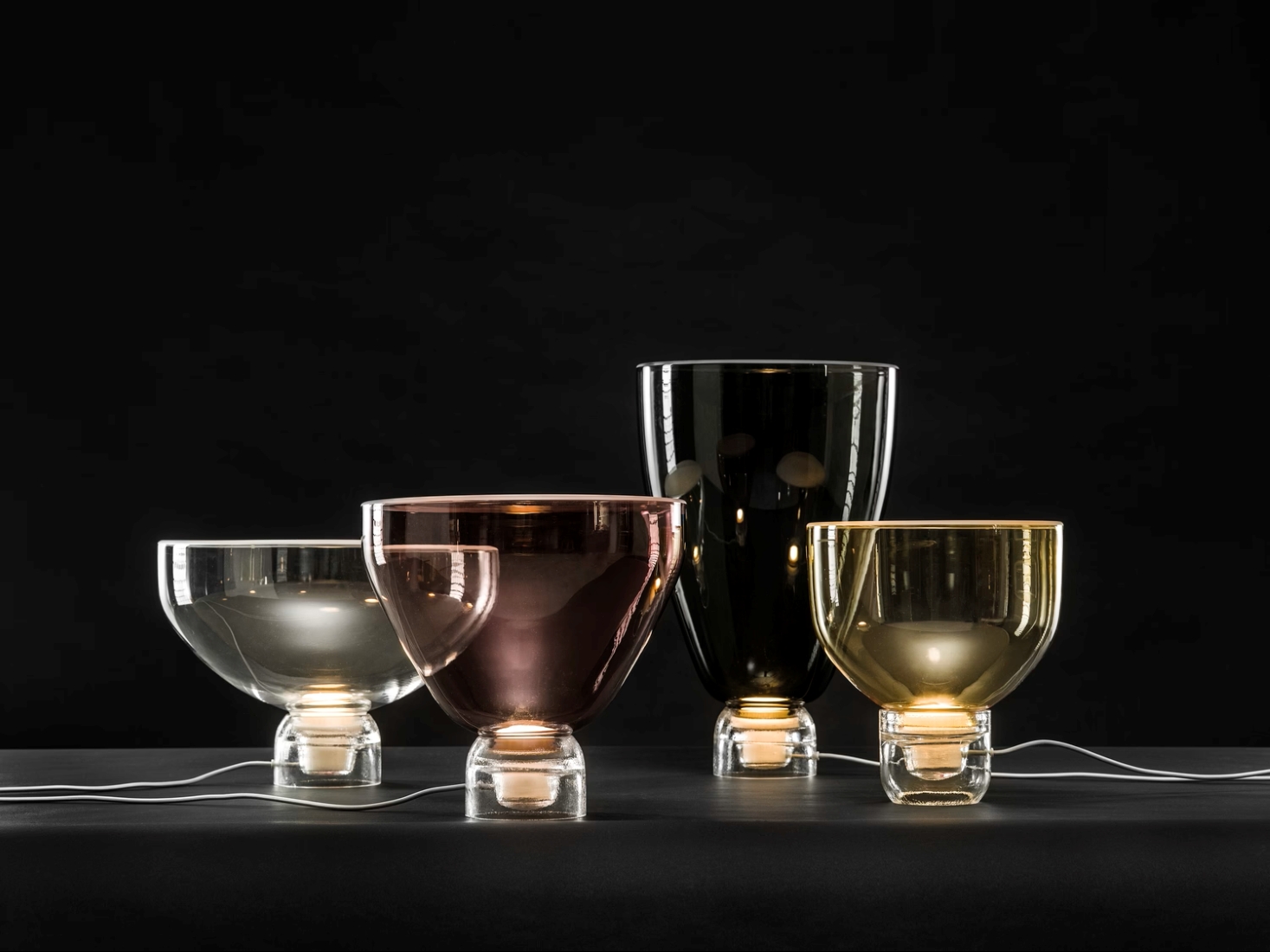 Brokis Lightline lamps in clear, violet, black, and amber glass glow luxuriously on a dark surface.