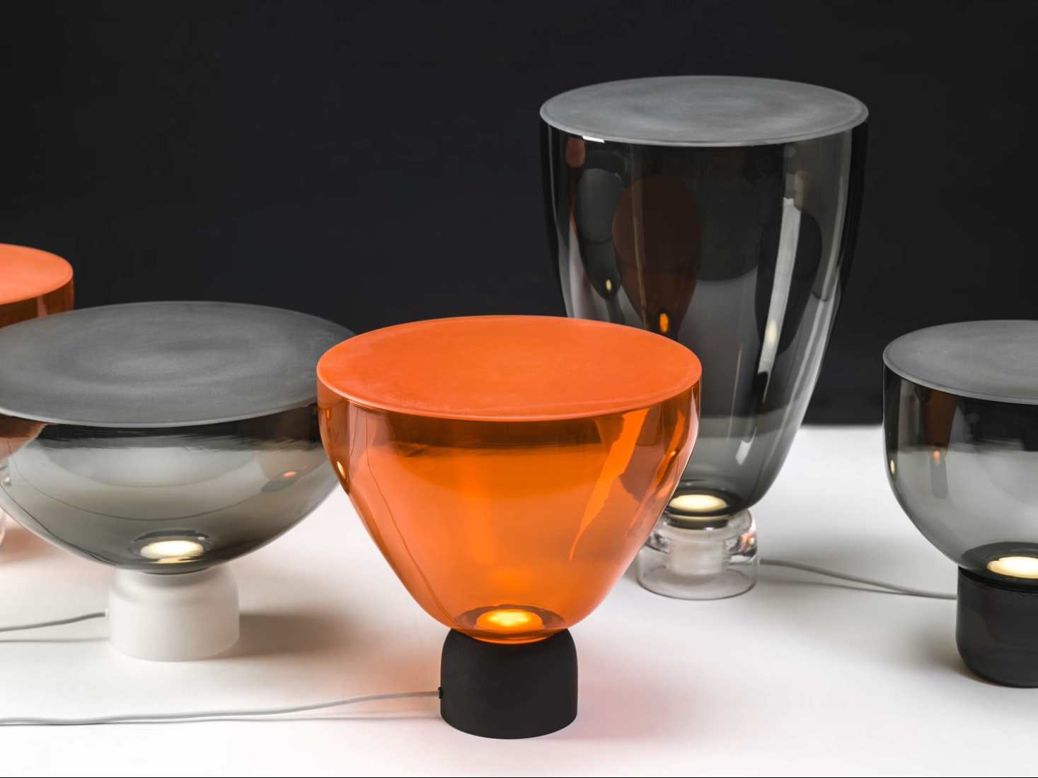 Five Brokis Lightline table lamps in grey and orange handblown glass glow on a white surface against a black backdrop.