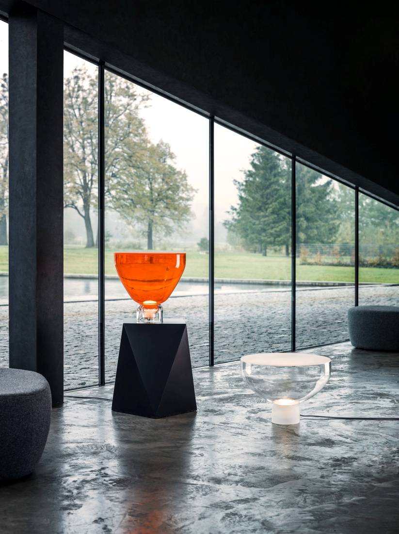 Sophisticated interior with glass walls framing misty greenery; orange Lightline S on plinth, sculptural Lightline L nearby.