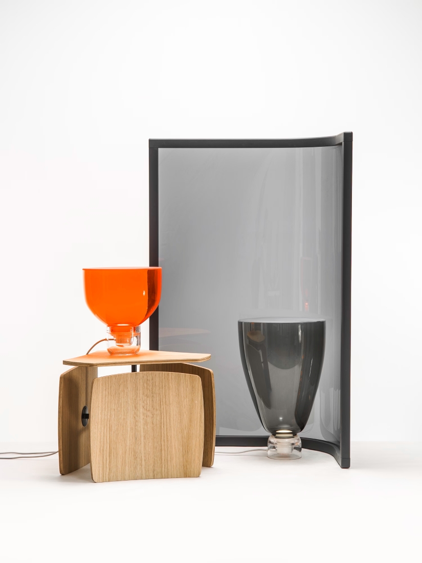 A modern still life with an orange Lightline S by Brokis on a wooden table, a Lightline L-H, and an arched grey screen.