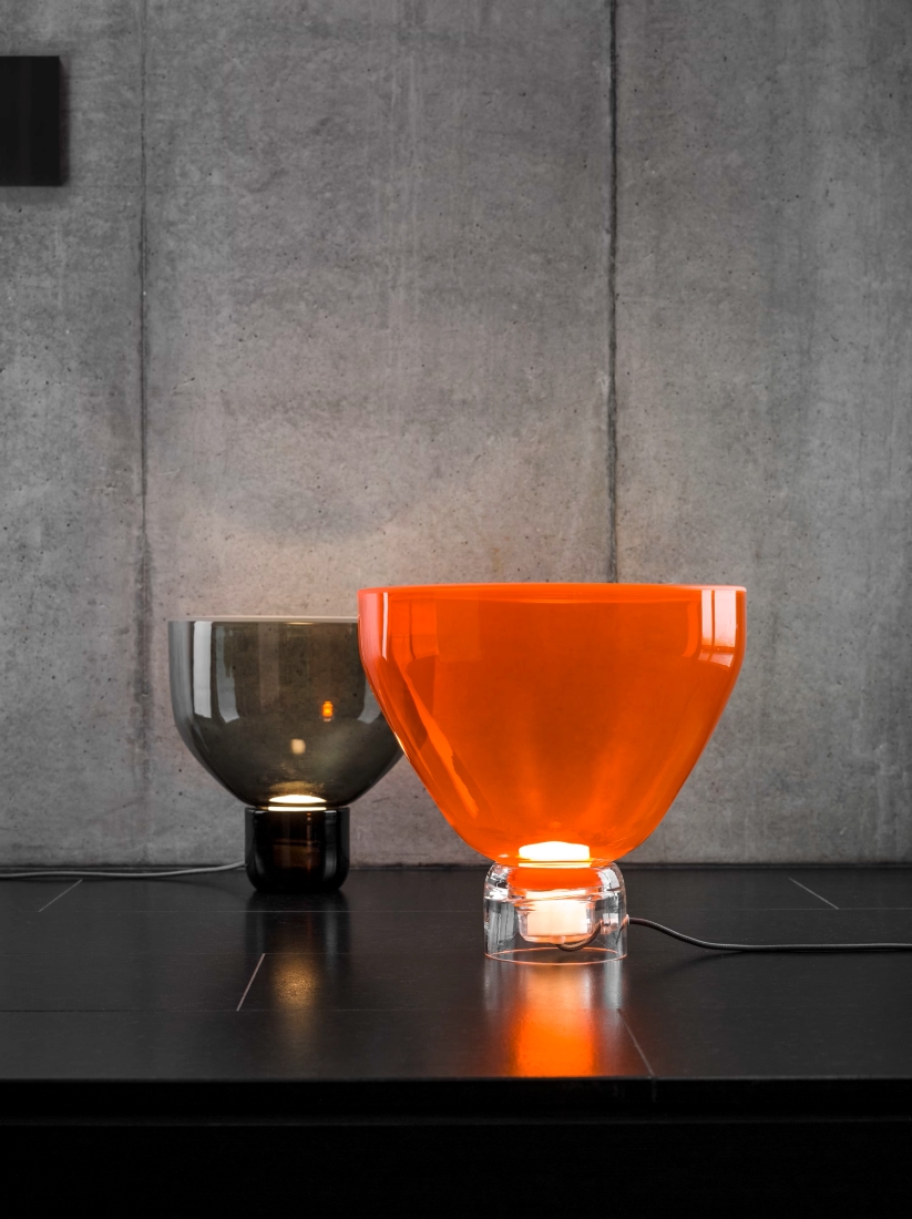 Two Brokis Lightline table lamps - one with a handblown orange glass shade, one in grey - accent a sleek black surface.