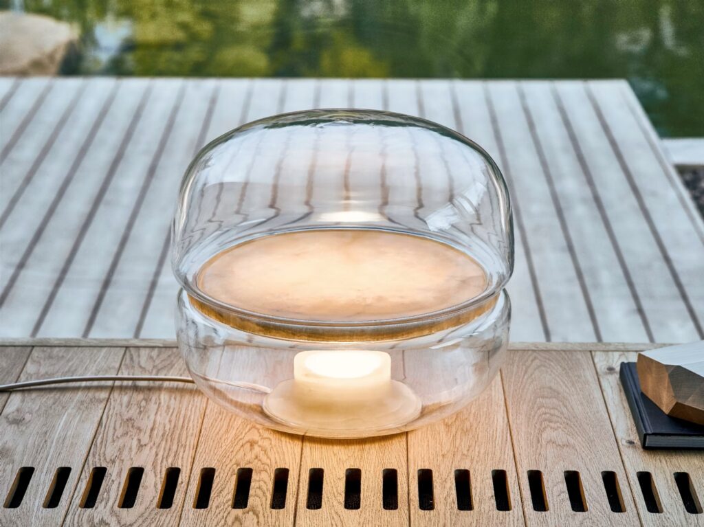 Brokis Macaron M lamp with a rounded crystal dome graces a wooden table by a serene deck and pond.