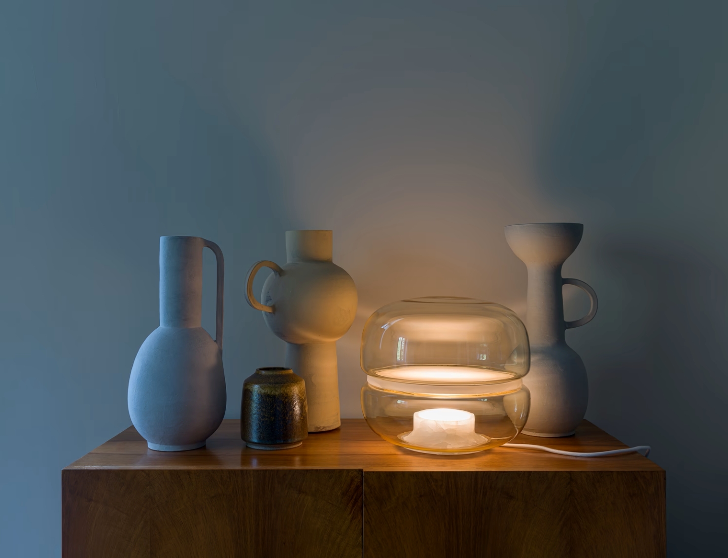 Sculptural ceramic vases cluster on wood, anchored by a Brokis Macaraon S lamp. Muted hues evoke serene modern luxury.