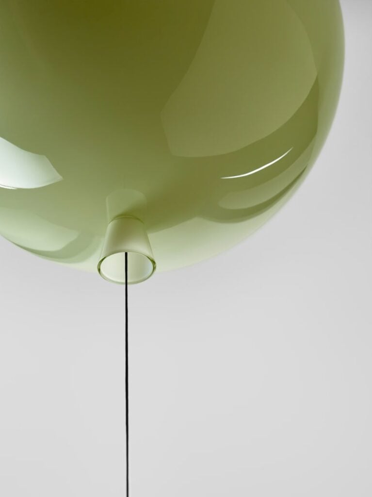 An olive green spherical Brokis Memory light with a cord, its smooth glass surface gleaming against a light grey backdrop.