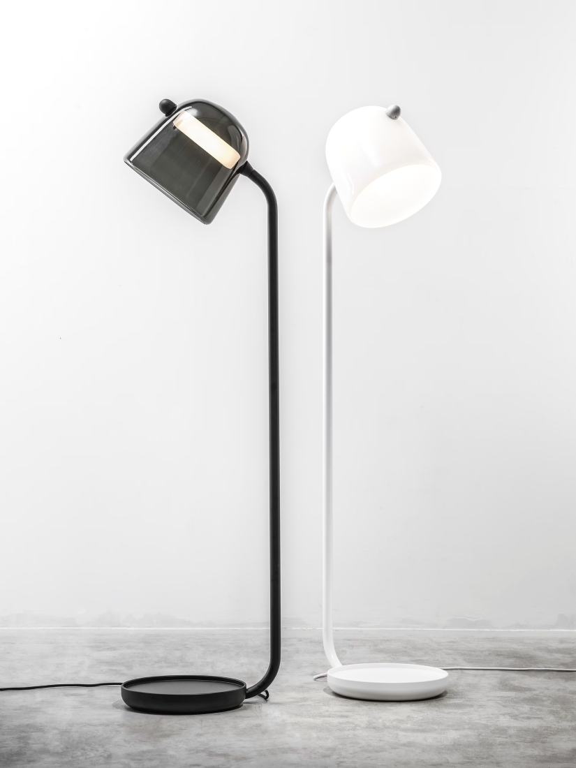 Two Brokis Mona Medium Floor lamps - one matte black, one white - stand elegantly side by side against a pristine white wall.