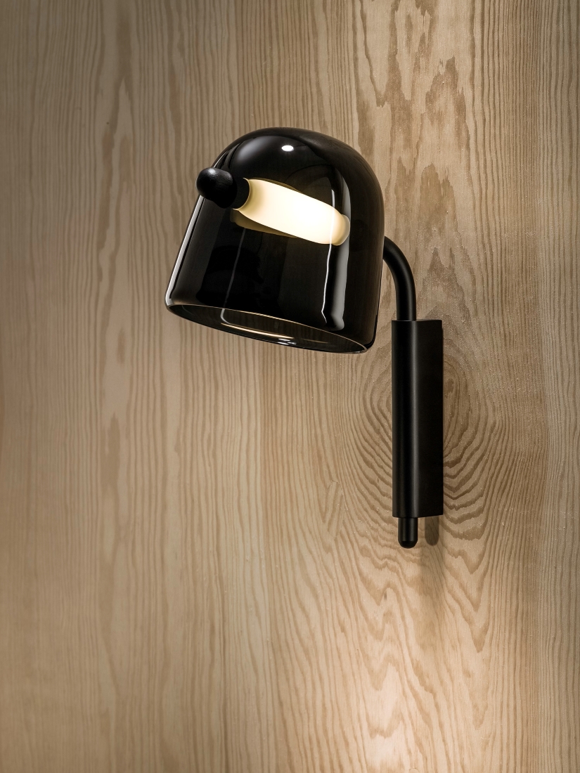 A black Brokis Mona Small Wall lamp with a glossy dome shade casts warm light against luxe wood grain and a minimalist arm.