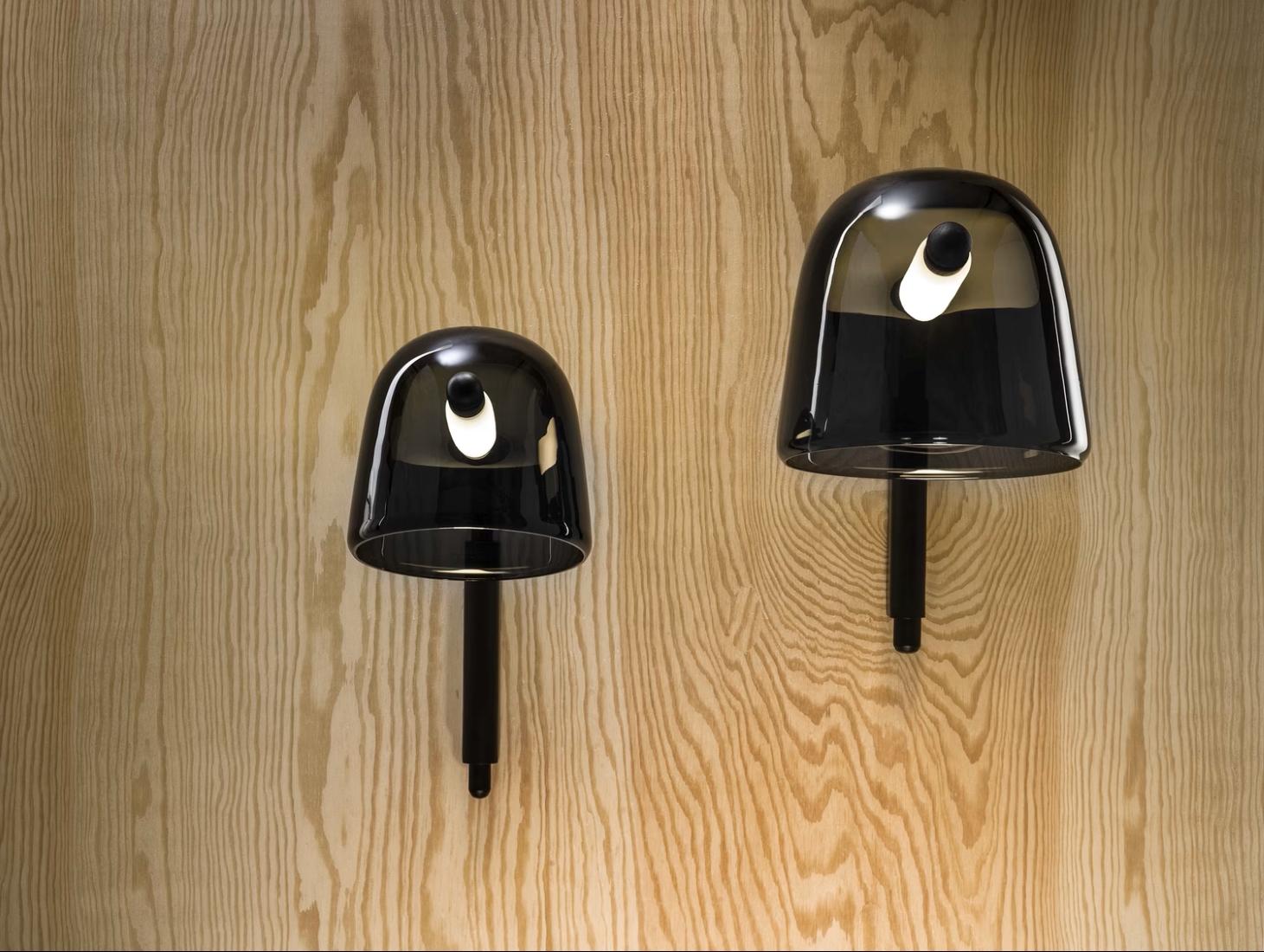 Two black Brokis Mona Wall lamps with glossy domes glow on a light wavy-grain wood wall, casting soft, elegant shadows.