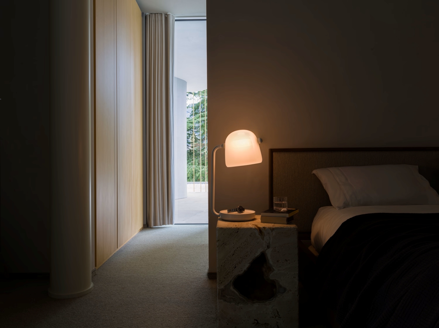 A dimly lit bedroom with plush bedding, a stone bedside table, Brokis Mona Small Table, books, and lush views.
