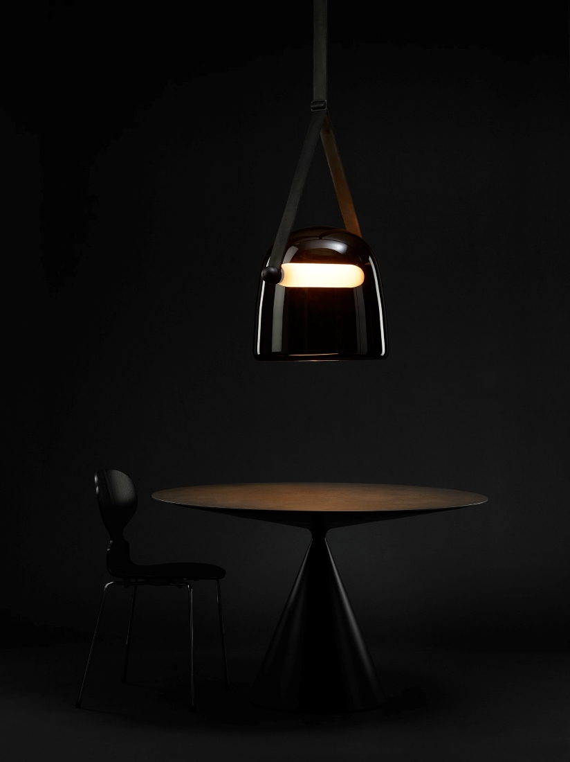 A round black table and chair sit beneath a Brokis Mona XL Pendant casting warm luxe light; moody black backdrop.
