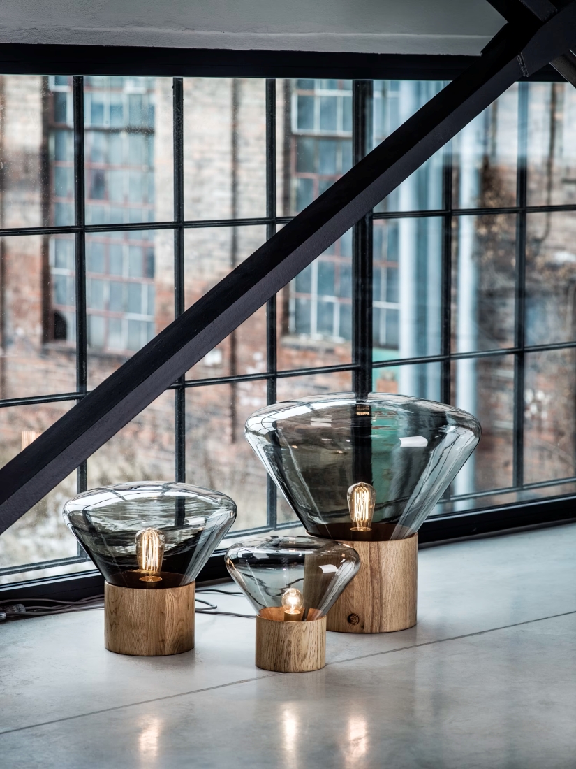 Brokis Muffins lamps with sculptural glass shades glow on wooden bases by an industrial window, exuding curated luxury.