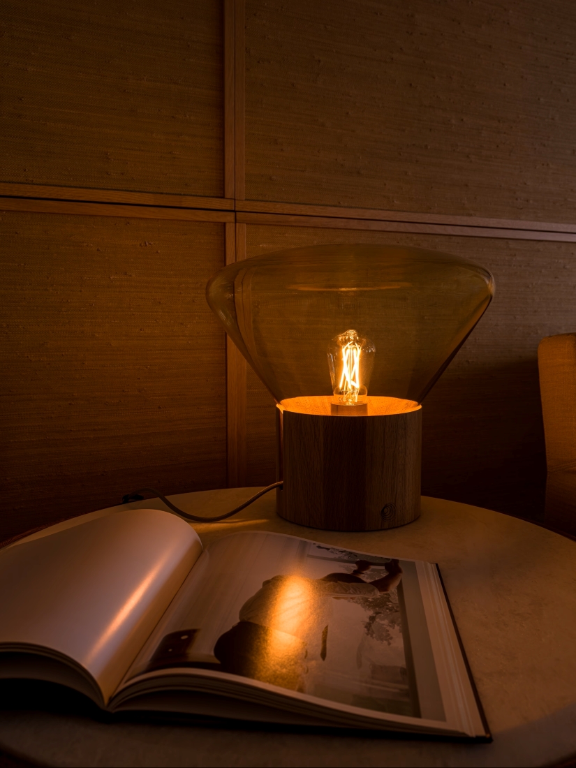 Brokis Muffins Wood 01 lamp glows atop a round table beside an art book in a warmly lit, wood-panelled interior.