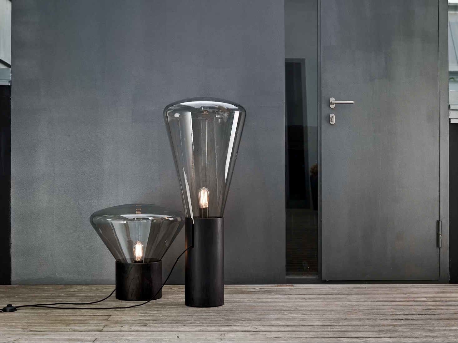 Two Brokis Muffins floor lamps with glass shades and black oak bases grace a wooden decking by a grey wall and glass door.