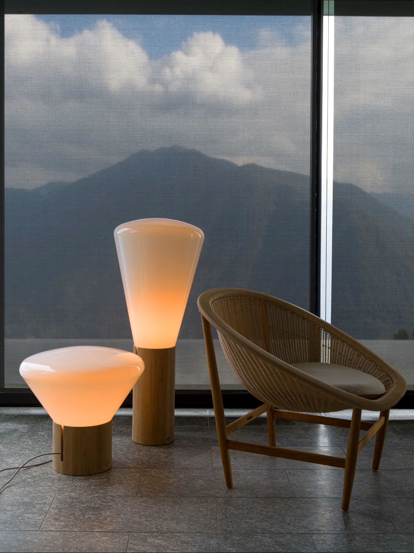 A modern room with panoramic windows reveals misty mountains. Brokis Muffins floor lamps and a wicker chair add luxe warmth.