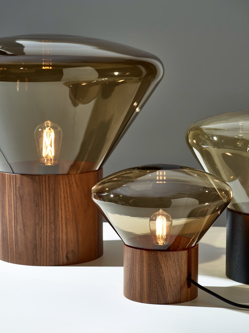 Three Brokis Muffins table lamps with hand-blown brown glass shades and wooden bases emit a luxe, warm glow on a white surface.