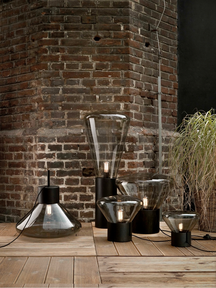 Brokis Muffins glass lamps with black oak bases glow on wood, set against rustic brick and airy greenery.