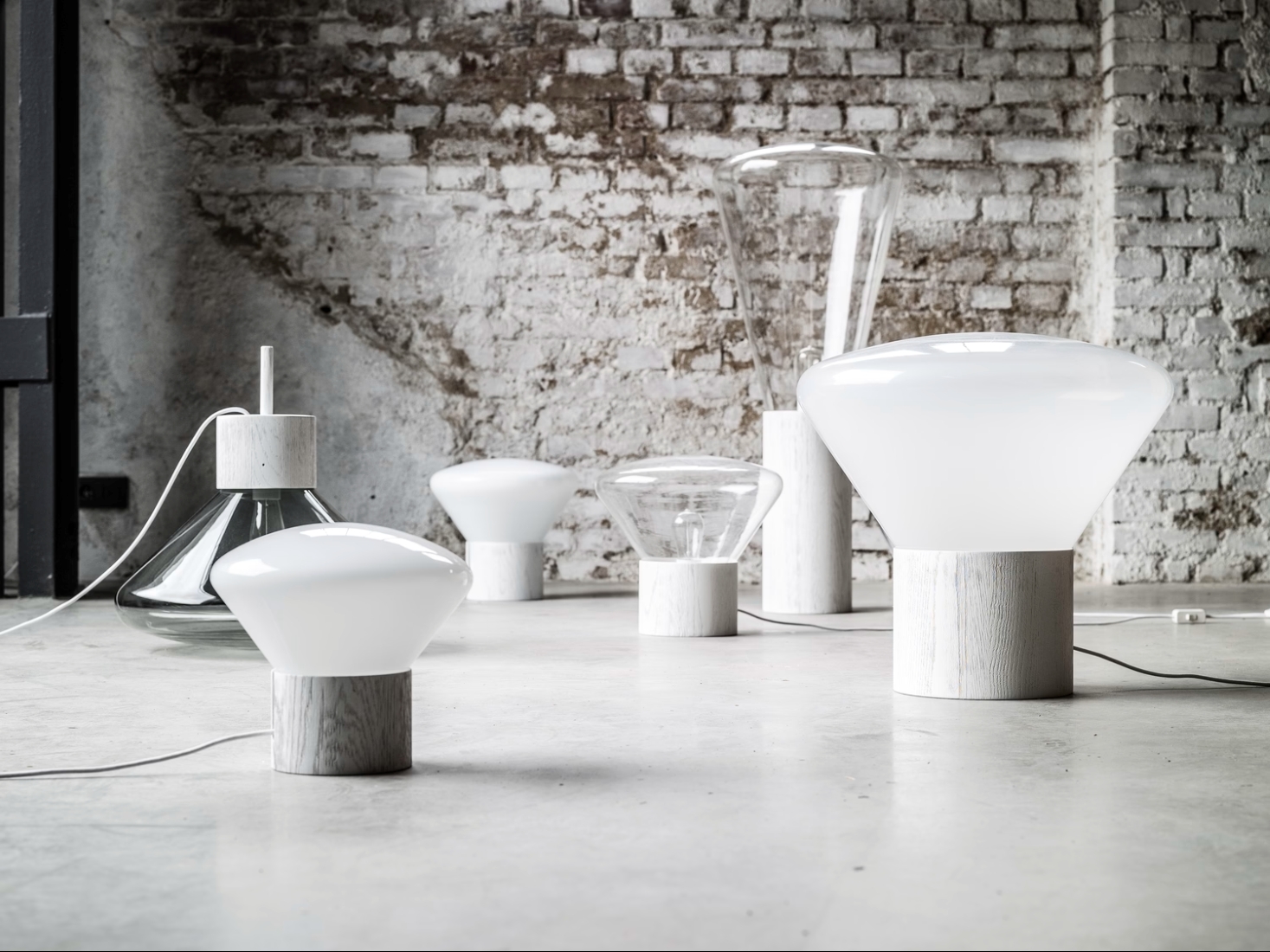 Brokis Muffins lamps with opaline glass glow on concrete, exuding minimalist luxury against a brick wall.