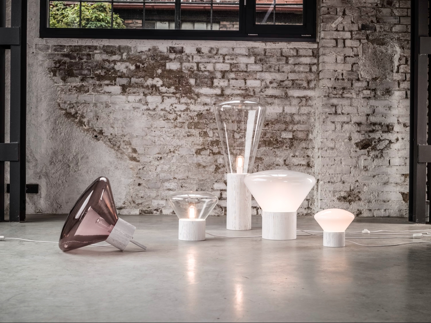 Brokis Muffins lamps in handblown white opaline glass glow softly on concrete, set against a luxe textured brick wall.