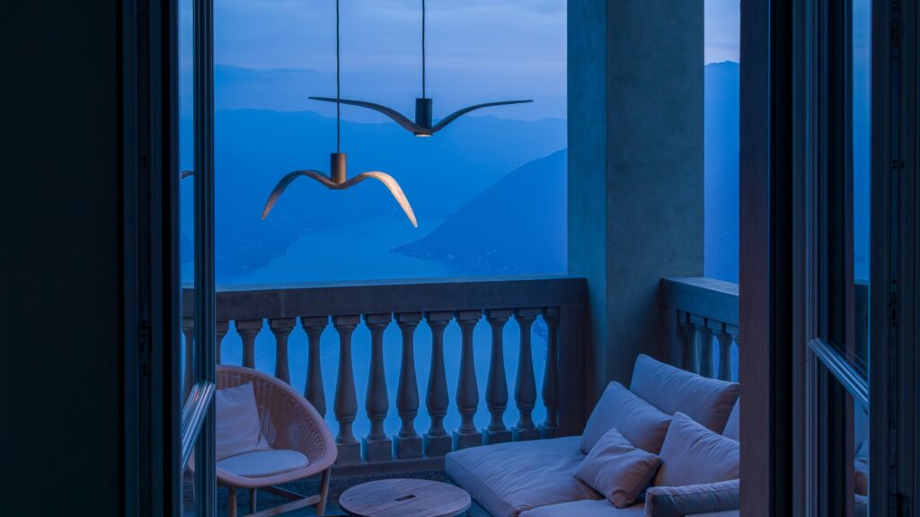 Brokis Night Birds pendants aglow in a dusk balcony with a plush sofa, round chair, and table overlooks a misty lake.