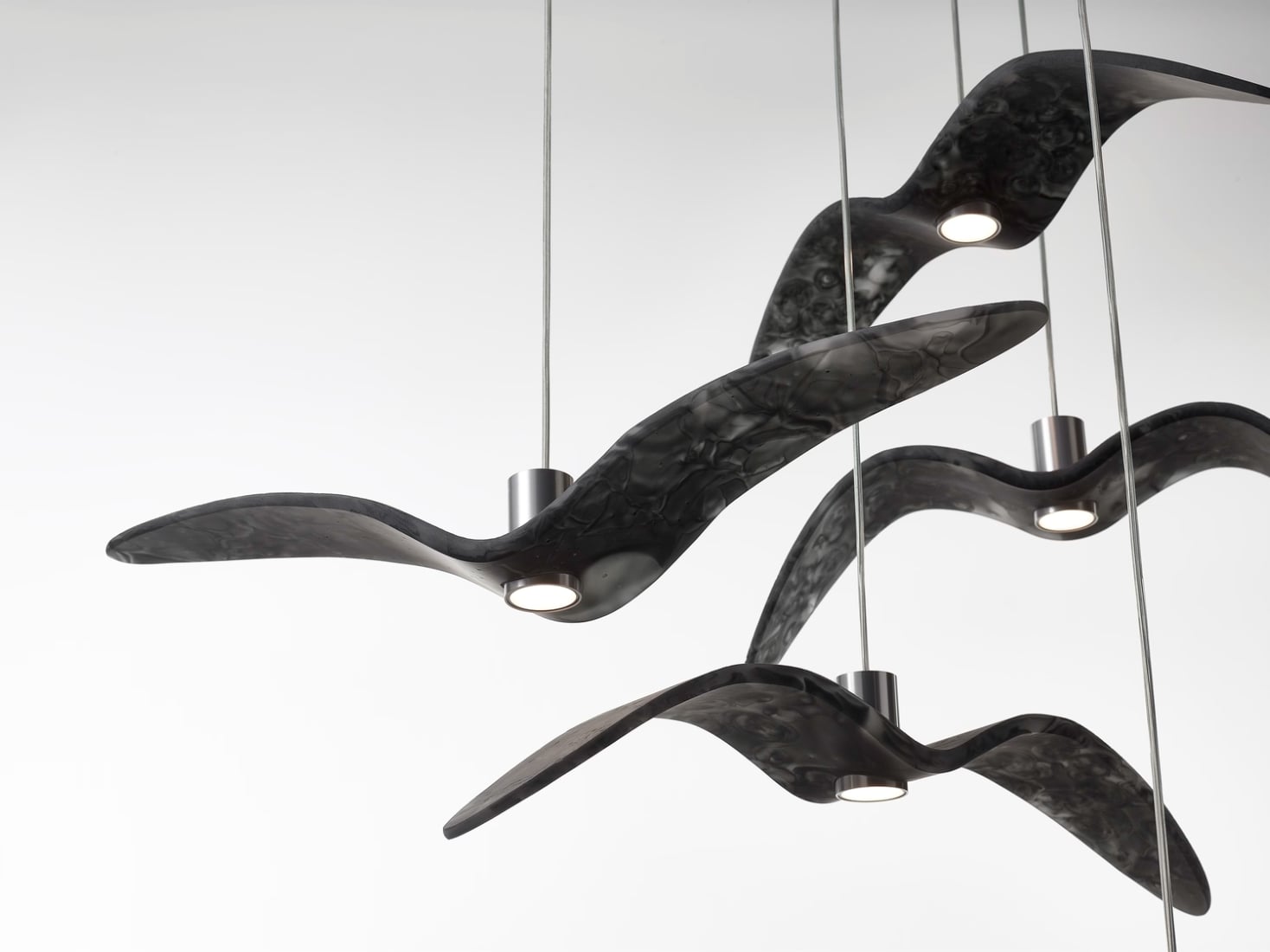 Brokis Night Birds pendants, handblown like abstract grey birds in flight, glow with circular bases against white.