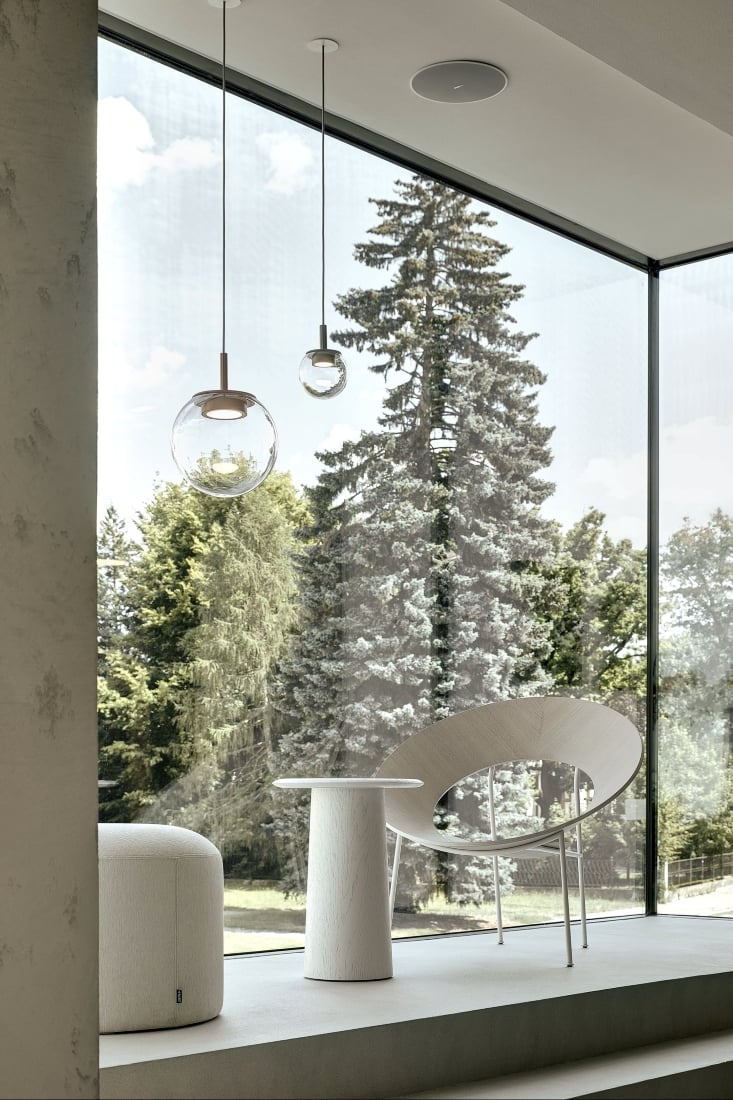 A luxurious minimalist space with Brokis Orbis pendants, rounded seating, and large windows framing lush pine views.