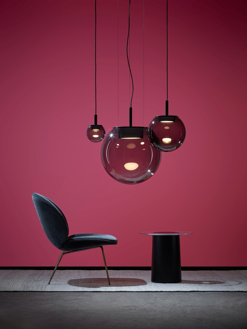 A dark velvet chair with gold legs, a glass-topped table, and three Brokis Orbis pendants on a plush grey rug.