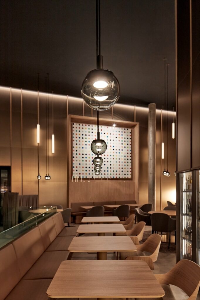 A modern restaurant with wooden tables, beige bench seating, and Brokis Orbis 150 pendants. Brass walls evoke refined warmth.