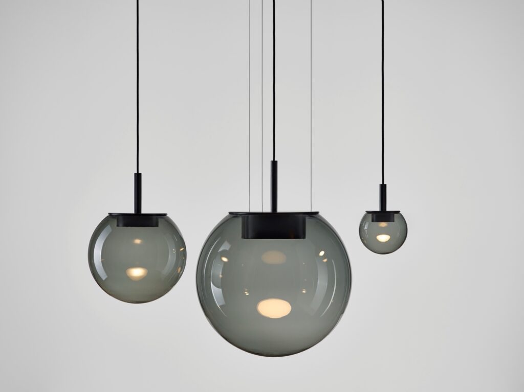 Three Brokis Orbis pendants with grey hand-blown glass globes and black accents glow against a pale grey backdrop.