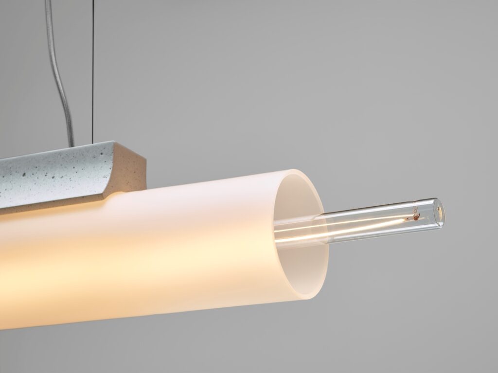 A close-up of a Brokis Overlay Chandelier L with handblown glass shade and glowing filament, against a soft grey backdrop.