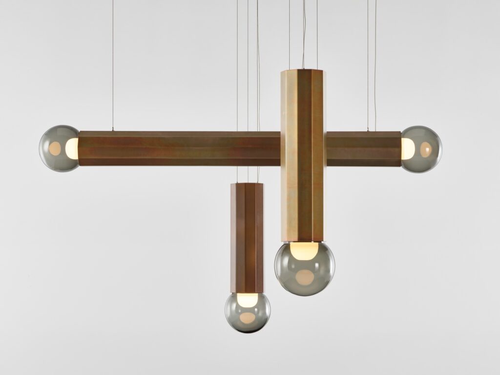 A Brokis Prisma chandelier with intersecting zinc rods and grey glass globes glowing warmly against a white background.