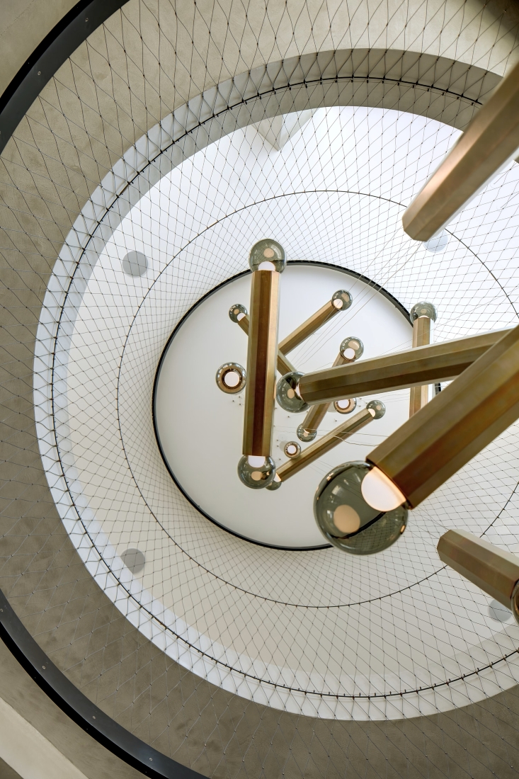 An upward view of a modern spiral staircase encircled by mesh, centring on gold rods and rings in a Brokis Prisma chandelier.