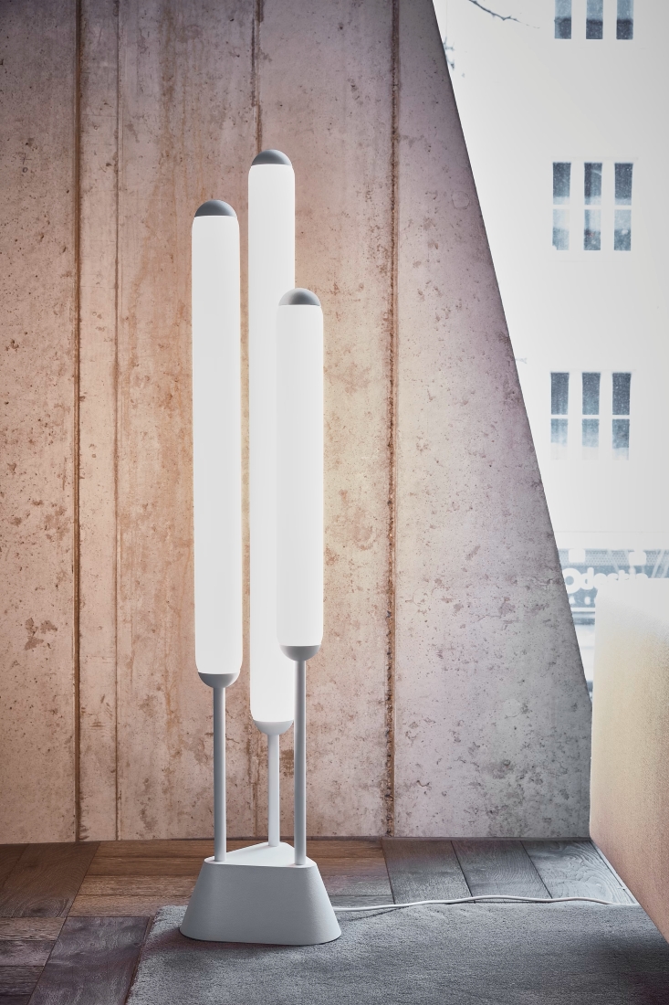 Brokis Puro Floor 3 Tubes 1350 with three white LED tubes on a grey base, set in a concrete room with dramatic windows.