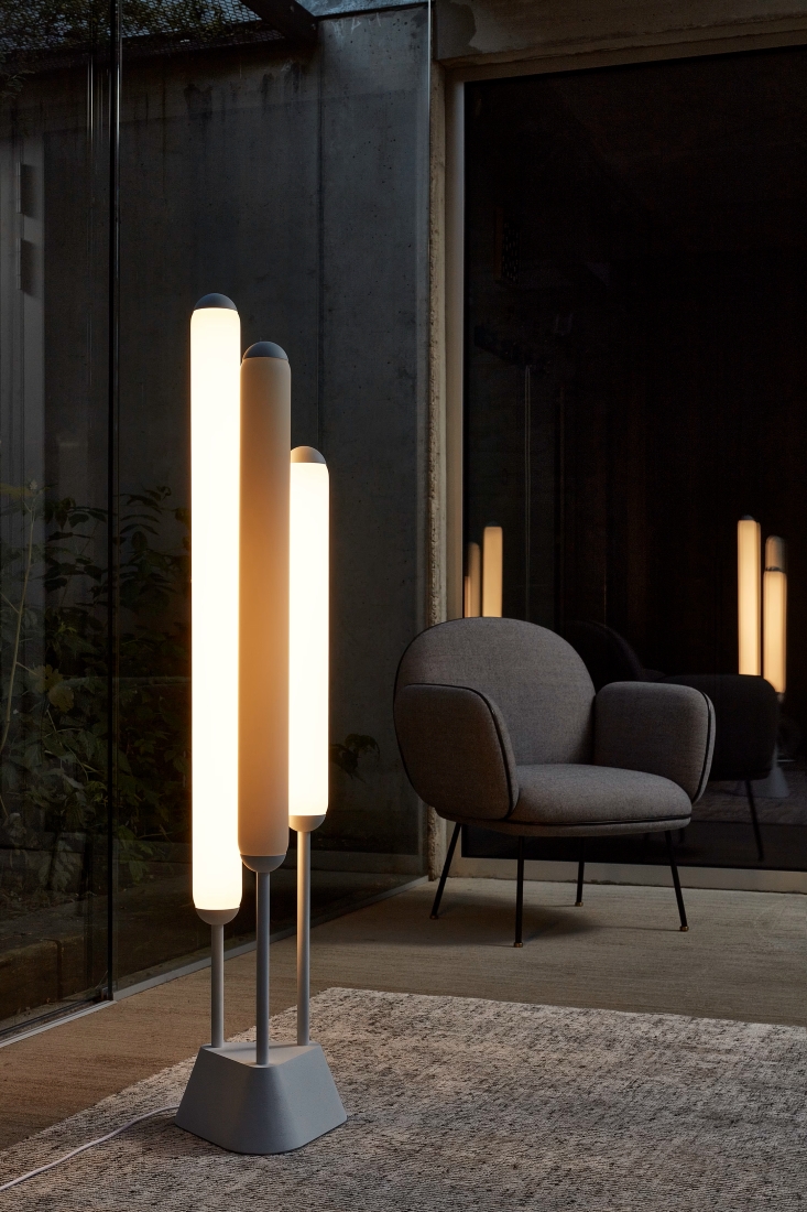 A modern room with Brokis Puro Floor 3 Tubes 1350 and plush grey armchair reflected in expansive glass windows.