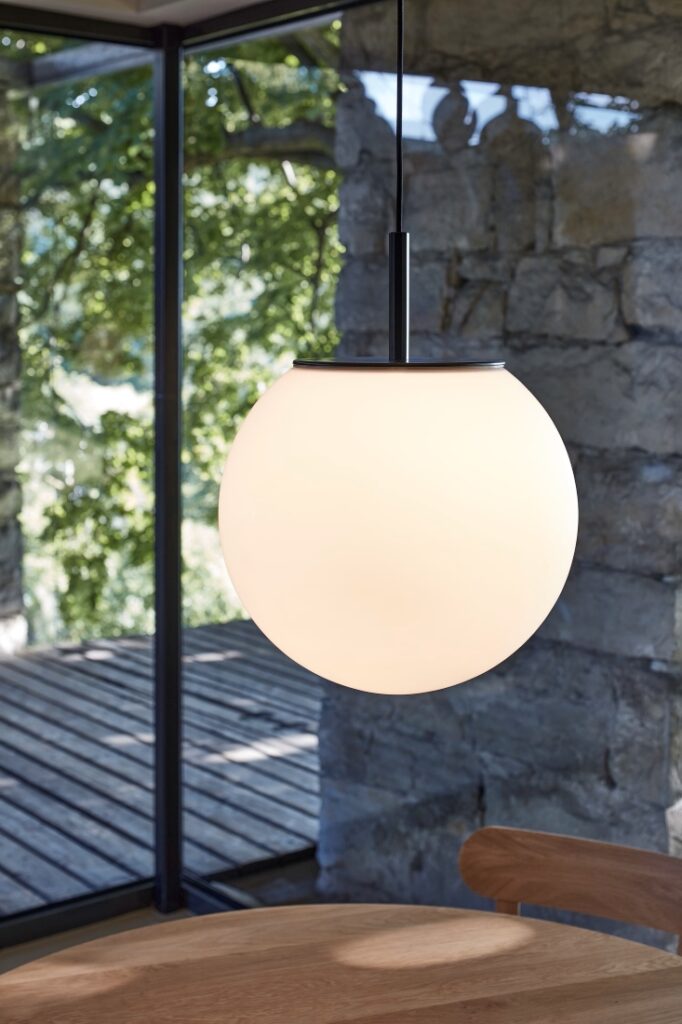 A Brokis Sfera 400 pendant floats over a wooden table, sunlight streaming through leafy windows.