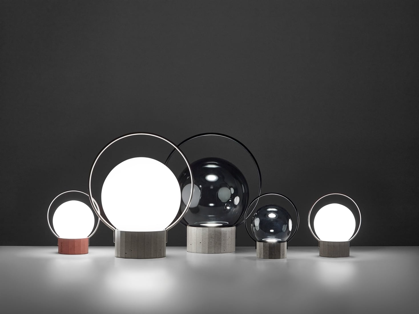 Five Brokis Sfera Portable table lamps in varying sizes glow atop a white surface, framed by a dramatic dark grey backdrop.