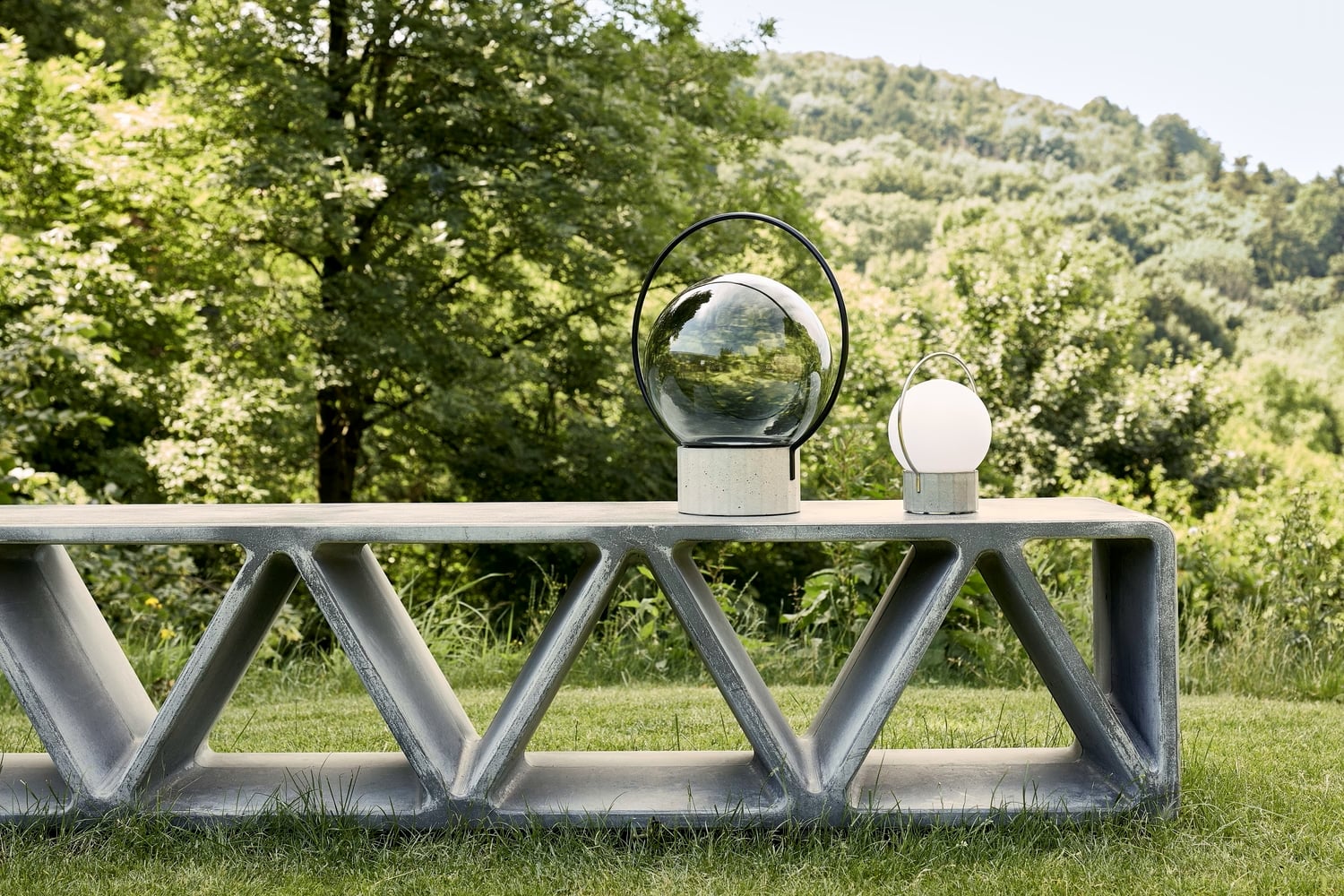 A sculptural concrete bench with triangular cut-outs holds two Brokis Sfera Portable lamps, set amid lush grass and trees.