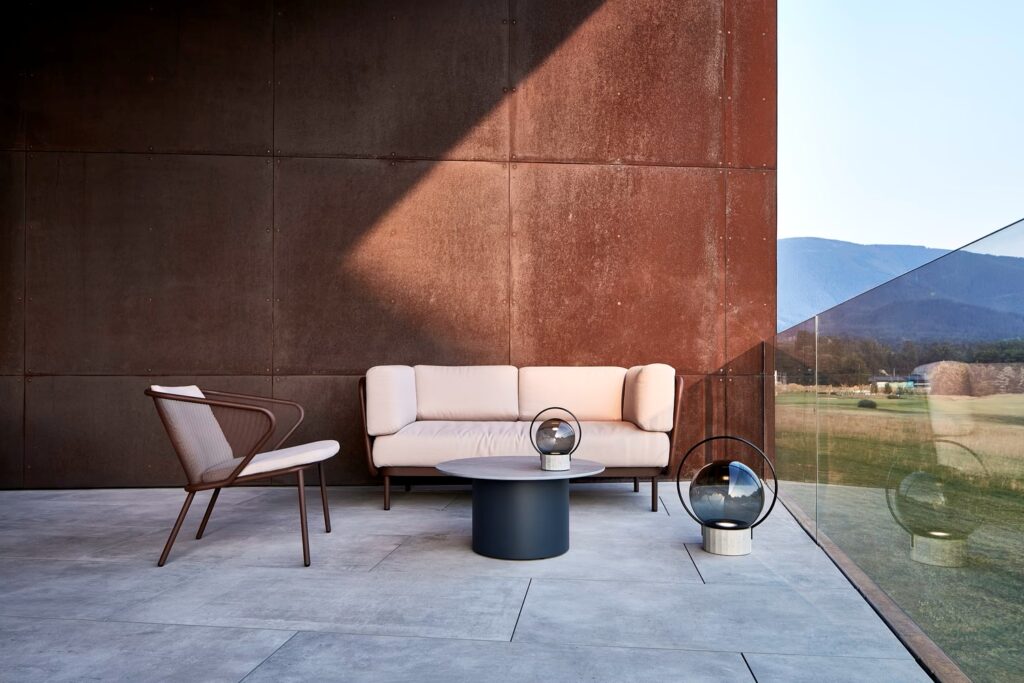 A modern patio with a white cushioned sofa, wood-accented chair, Brokis Sfera Portable lamps, and panoramic mountain views.