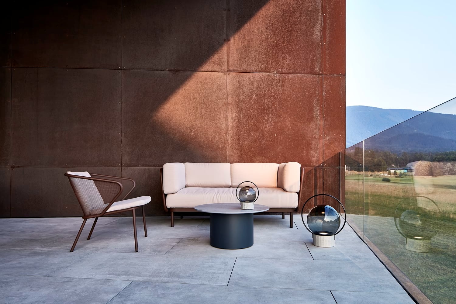 A modern patio with a white cushioned sofa, wood-accented chair, Brokis Sfera Portable lamps, and panoramic mountain views.