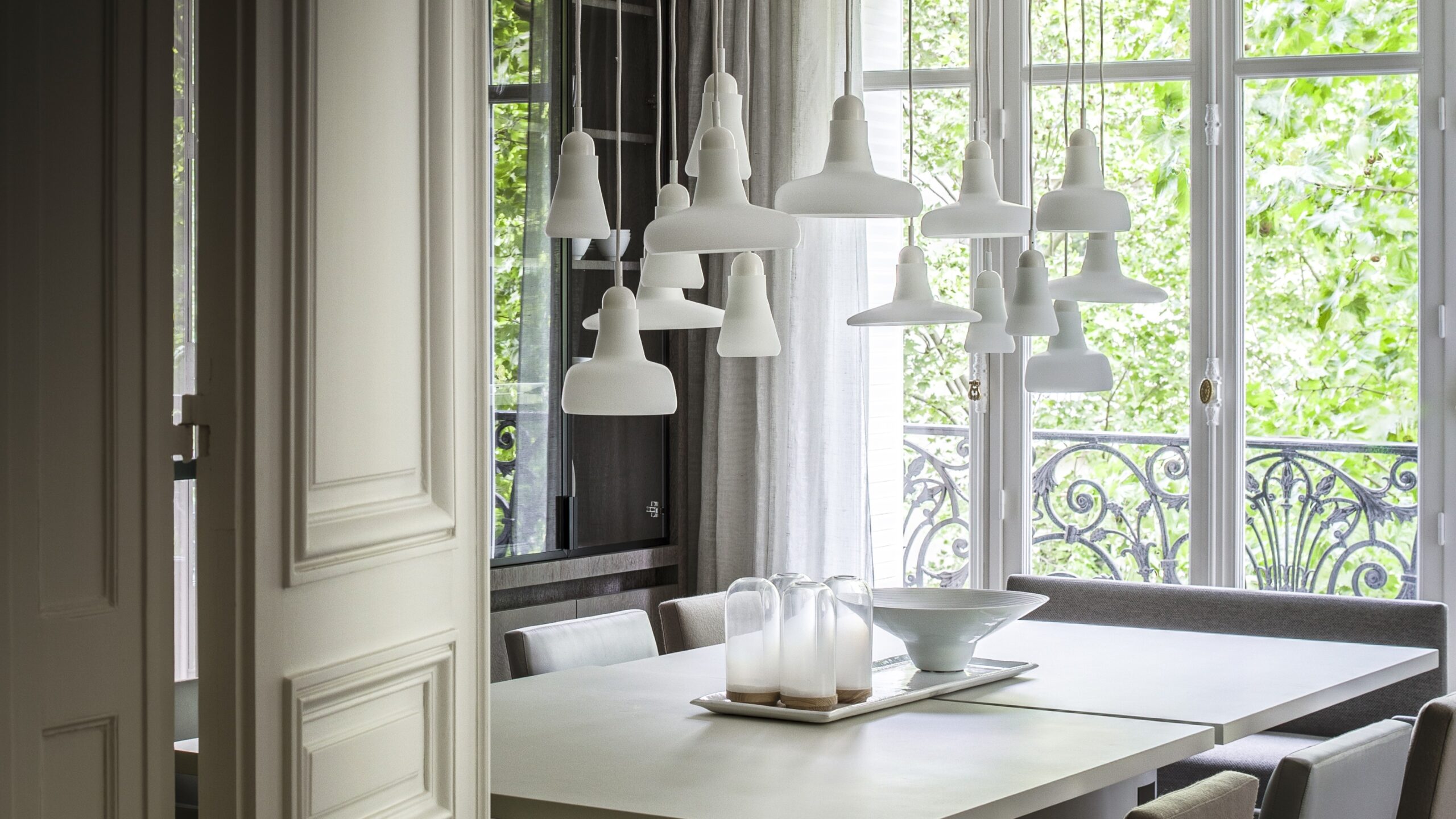 Brokis Shadows pendants above a long table, elegant grey chairs, and lush greenery visible outside.