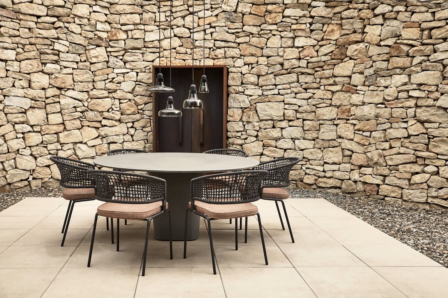 A table and six chairs, beneath Brokis Shadows Exterior pendants; stone walls complete this serene, elevated courtyard.
