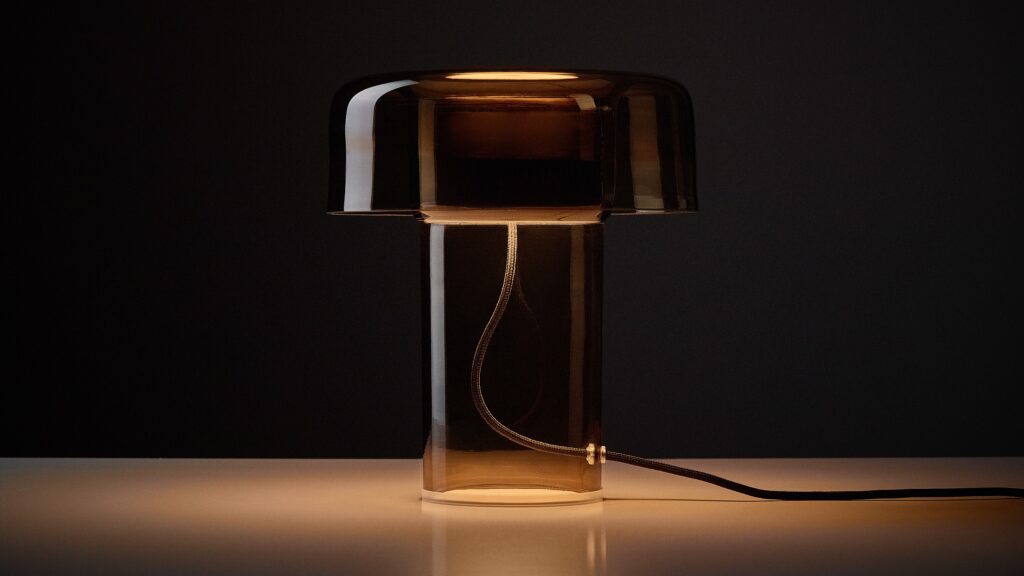 Brokis Spectra table lamp in hand-blown brown glass emits a warm glow atop a white surface, set against a dark backdrop.